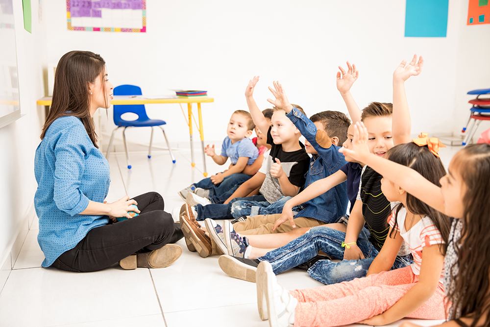 Professora e alunos do ensino infantil sentados no chão conversando e fazendo atividades para explorar e avaliar os campos de experiência.