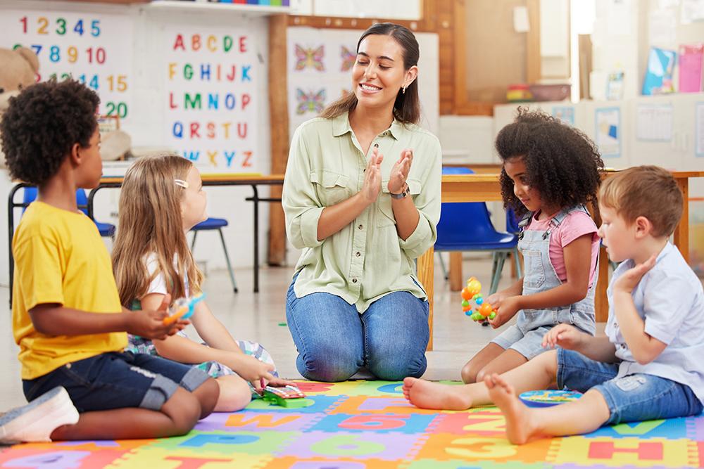 Professora de ensino infantil sentada em tapete ao lado de quatro alunos conversando e brincando.