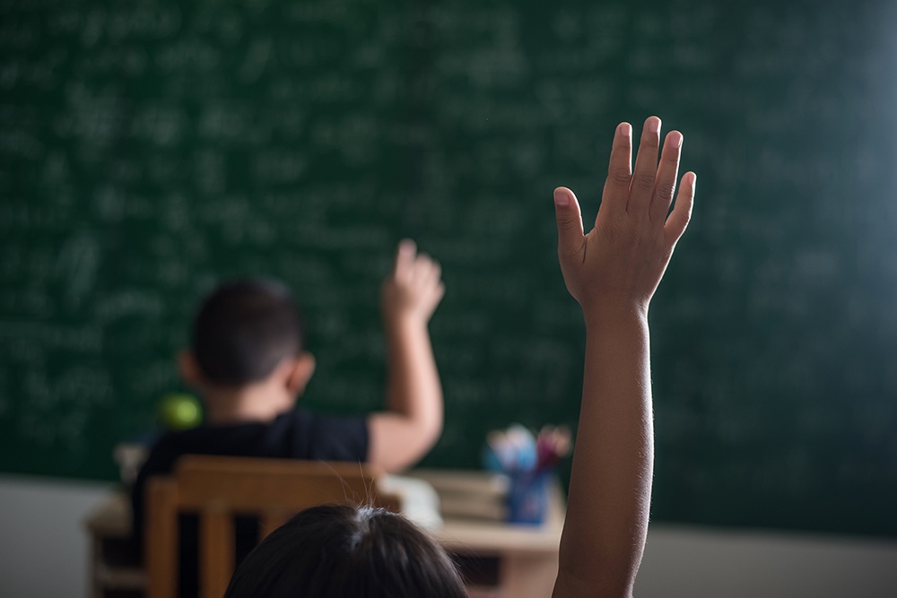 Crianças em sala de aula com as mãos levantadas para fazer perguntar para o professor.