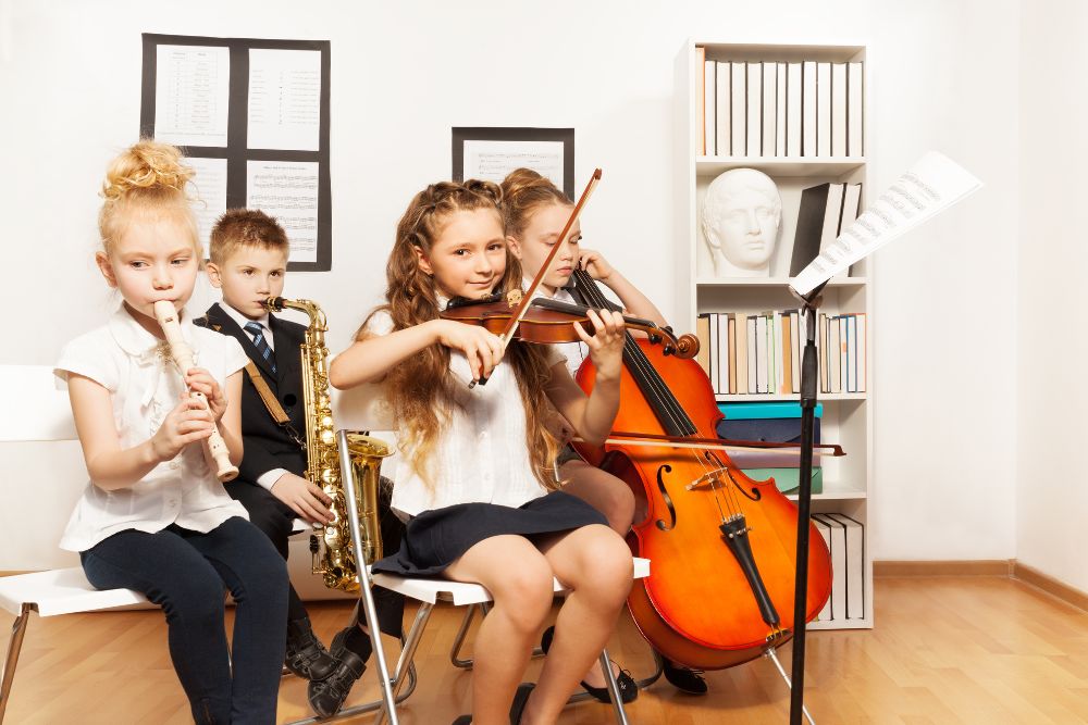 Um grupo de quatro crianças, com diferentes instrumentos musicais (flauta, saxofone, violino e violoncelo), tocando em uma sala de aula, com partituras nas estantes ao fundo.