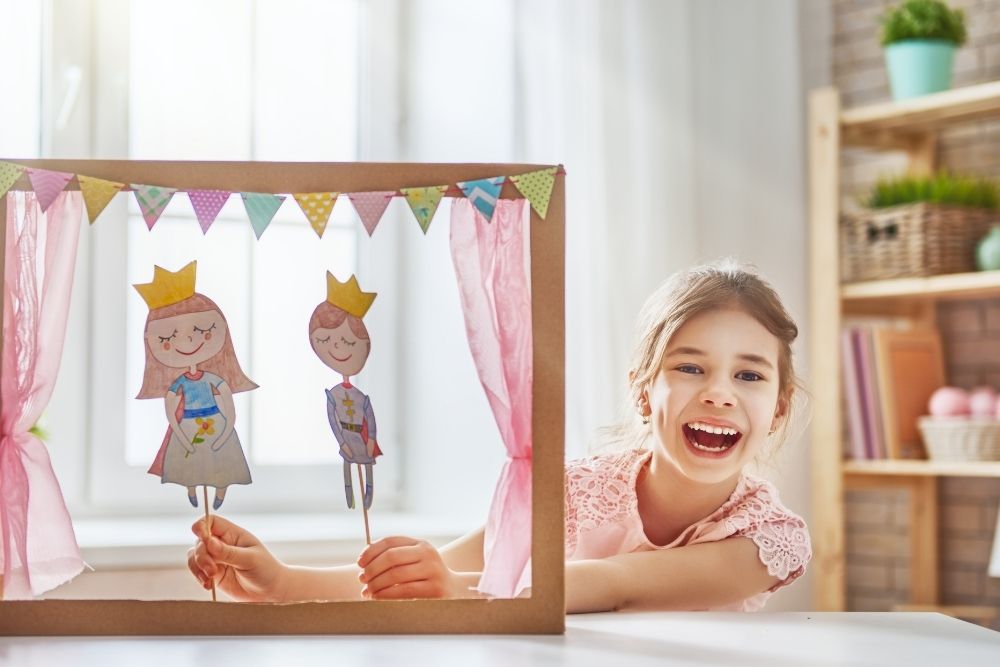 Uma menina sorridente, de cabelo preso, segurando fantoches de papel que representam personagens de um teatro infantil em miniatura, com um cenário colorido ao fundo.