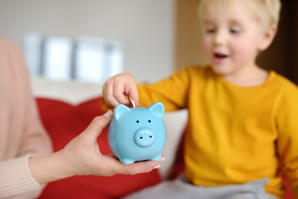 menino colocando moeda em cofrinho azul, aprendendo sobre poupança.