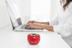 timer de cozinha em formato de tomate sobre uma mesa enquanto uma mulher estuda no laptop.